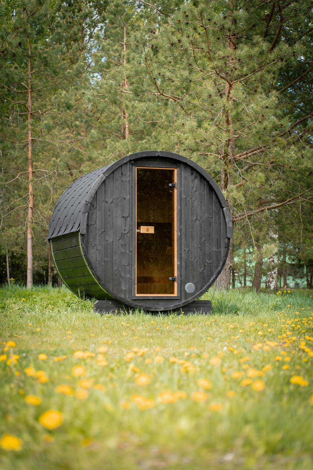 PlusLife Cove Barrel Sauna in black-stained timber, end on, with a tall narrow glass door showing the timber interior, standing in grass among pine trees