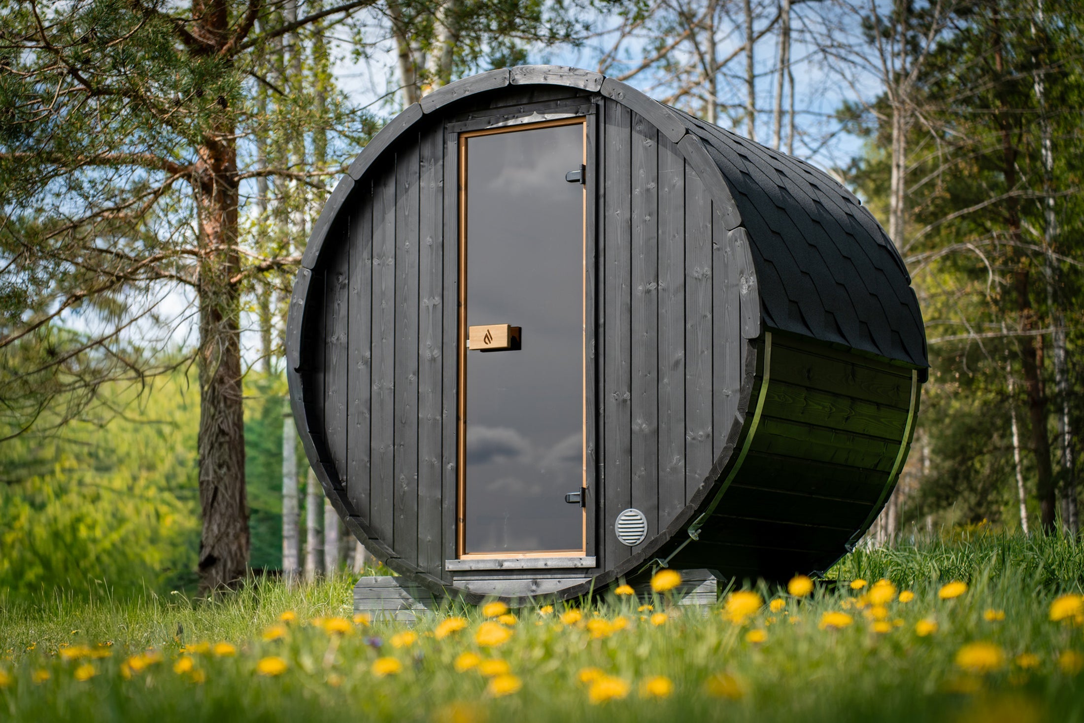Cove Barrel sauna in black-stained timber with a black shingle roof, a tall narrow tinted-glass door and timber cradle feet, in a garden